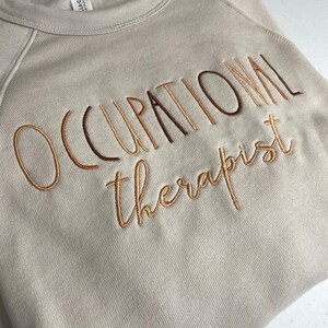 Może przedstawiać: Beżowa bluza z haftem "Occupational therapist" wykonanym brązową i złotą nicią.