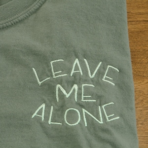 Può includere: Una maglietta verde con la frase "Leave Me Alone" ricamata in bianco sul davanti.