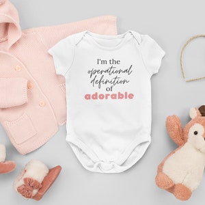 Peut inclure: Combinaison blanche pour bébé à manches courtes avec le texte "I'm the operational definition of adorable" en lettres noires et roses.