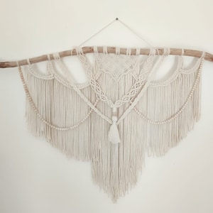 May include: A white macrame wall hanging with a wooden branch at the top and a tassel hanging from the bottom. The macrame is made with a variety of knots and patterns, and it has a bohemian style.