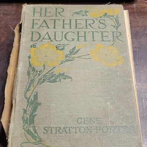 May include: A vintage book titled "Her Father's Daughter" by Gene Stratton-Porter. The cover features a floral design with yellow flowers and green leaves on a textured, light brown background.