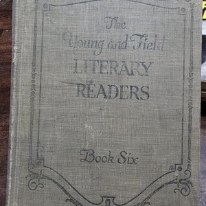 Puede incluir: Un libro antiguo titulado "The Young and Field Literary Readers, Book Six". La portada es de color verde apagado con adornos y letras decorativas en negro. El libro parece desgastado, con una superficie texturizada.