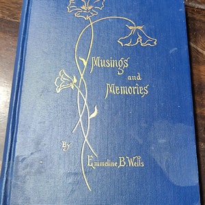 May include: A blue book with gold lettering that reads "Musings and Memories" by "Emmeline B. Wells". The cover features a decorative design of stylized flowers.