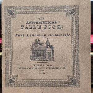 Op de afbeelding: Een bruinachtige boekcover met de titel "The Arithmetical Table Book" en de ondertitel "First Lessons in Arithmetic". De cover heeft zwarte tekst en een kleine illustratie van een gebouw. Gedrukt in Newark, N.J., in 1844.