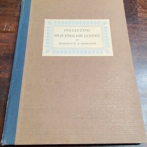 Może przedstawiać: Brązowa książka z niebieskim grzbietem. Okładka ma białą etykietę z niebieską obwódką i tekstem "COLLECTING OLD ENGLISH LUSTRE by JEANNETTE R. HODGDON".