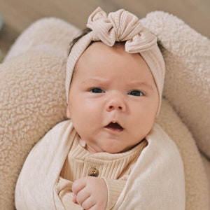 May include: A baby wearing a beige knitted headband with a bow on top. The headband is tied around the baby's head and the bow is centered on top of the baby's forehead. The baby is wearing a beige knitted onesie and is looking directly at the camera.