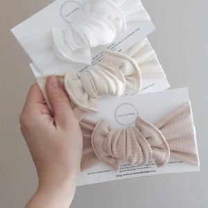May include: Three knotted headbands in different shades of beige. The headbands are made of a soft, stretchy fabric. The text "Emi & Jo Baby" is visible on the packaging.