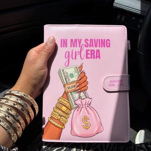 May include: A light pink rectangular binder featuring the text "IN MY SAVING girl ERA" in pink and white. The cover displays an illustration of a hand holding a stack of US dollar bills and a pink money bag with a dollar sign, plus a small tab labeled "SAVING GIRL ERA".