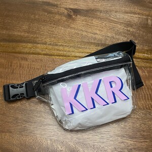 May include: Clear plastic fanny pack with a black adjustable strap and a zipper closure. The fanny pack has a white lining and a pink and blue "KKR" logo.