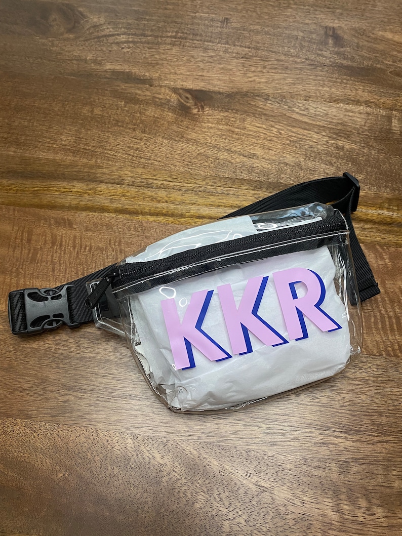 May include: Clear plastic fanny pack with a black adjustable strap and a zipper closure. The fanny pack has a white lining and a pink and blue "KKR" logo.