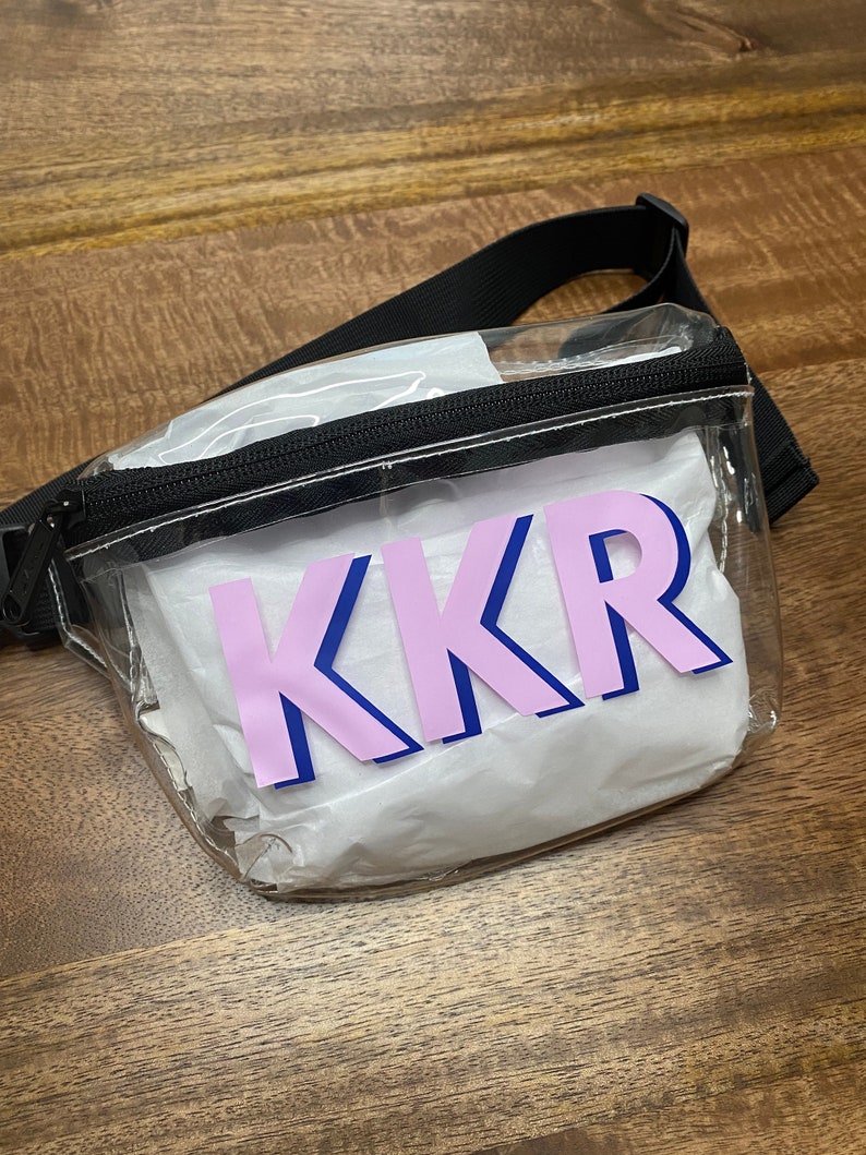 May include: Clear plastic fanny pack with a black adjustable strap and a pink and blue "KKR" design.