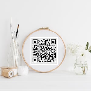 May include: A black and white QR code is displayed in a wooden embroidery hoop. The hoop is on a white surface with a vase of white flowers, spools of thread, and paintbrushes in a glass jar.