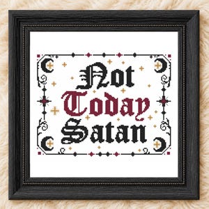 Op de afbeelding: Een ingelijst kruissteek borduurwerk met de tekst Not Today Satan in zwarte en donkerrode gotische letters. Het ontwerp is omgeven door een decoratieve rand met zwarte halve manen, kleine gouden sterren en donkerrode accenten op een witte ondergrond.
