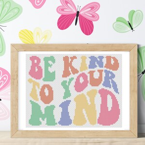 Peut inclure: Une broderie au point de croix encadrée avec un fond blanc et un texte coloré qui dit "Be Kind To Your Mind". Le cadre est en bois brun clair et il y a des papillons en arrière-plan.