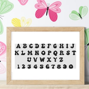 Può includere: Un ricamo a punto croce incorniciato che presenta l'alfabeto e i numeri in nero su uno sfondo bianco. La cornice è in legno marrone ed è circondata da uno sfondo bianco con illustrazioni di farfalle colorate.