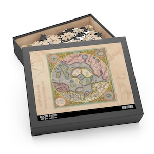 Puede incluir: Caja de rompecabezas negra abierta que revela un mapa antiguo del Ártico. El mapa circular presenta una rosa de los vientos y bordes decorativos. La caja está etiquetada como "16x20 Puzzle 500 pcs Ages 9+". Mide 40,64 cm x 50,8 cm.