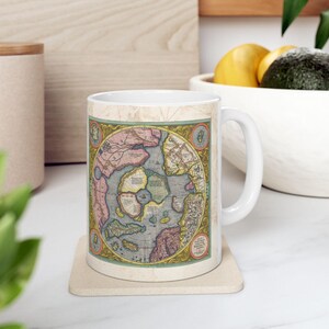 Puede incluir: Taza de cerámica blanca con un mapa vintage del Ártico impreso en ella. El mapa está en tonos de azul, verde, amarillo y rosa. La taza tiene un asa en el lado derecho.