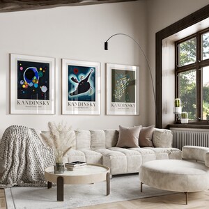 May include: Three framed art prints with abstract designs by artist Wassily Kandinsky. The prints are in black frames and feature the artist's name and the title of the artwork. The prints are hanging on a white wall in a living room setting with a couch, coffee table, and floor lamp.
