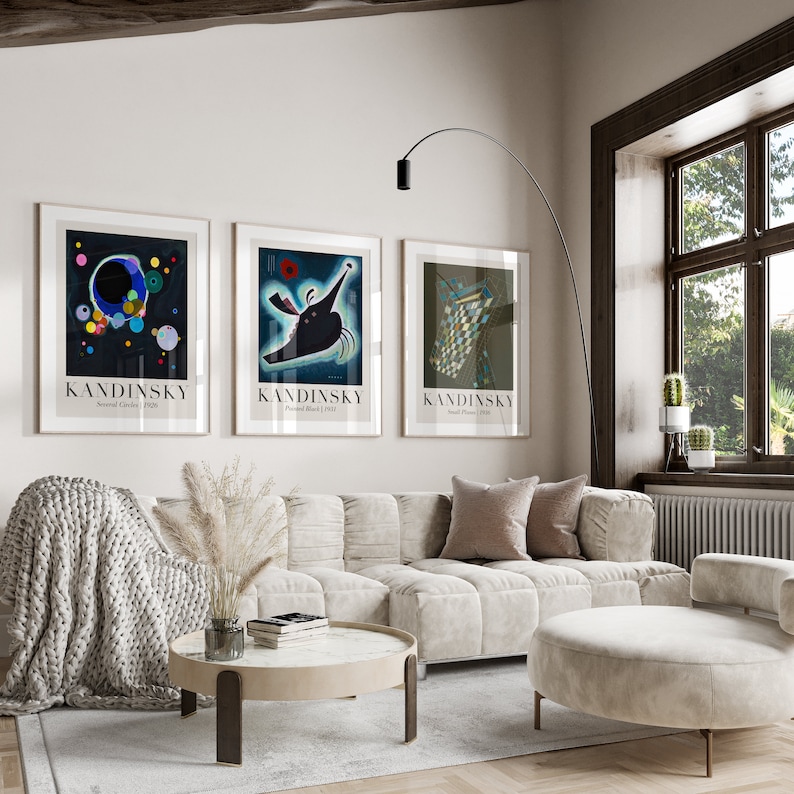 May include: Three framed art prints with abstract designs by artist Wassily Kandinsky. The prints are in black frames and feature the artist's name and the title of the artwork. The prints are hanging on a white wall in a living room setting with a couch, coffee table, and floor lamp.
