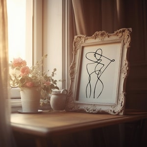 K&ouml;nnte beinhalten: Ein wei&szlig; gerahmtes Bild mit einer schwarzen Linienzeichnung einer Person mit Hut. Das Bild steht auf einem Holztisch mit einer Vase mit rosa Blumen und einem kleinen Keramikgef&auml;&szlig;.