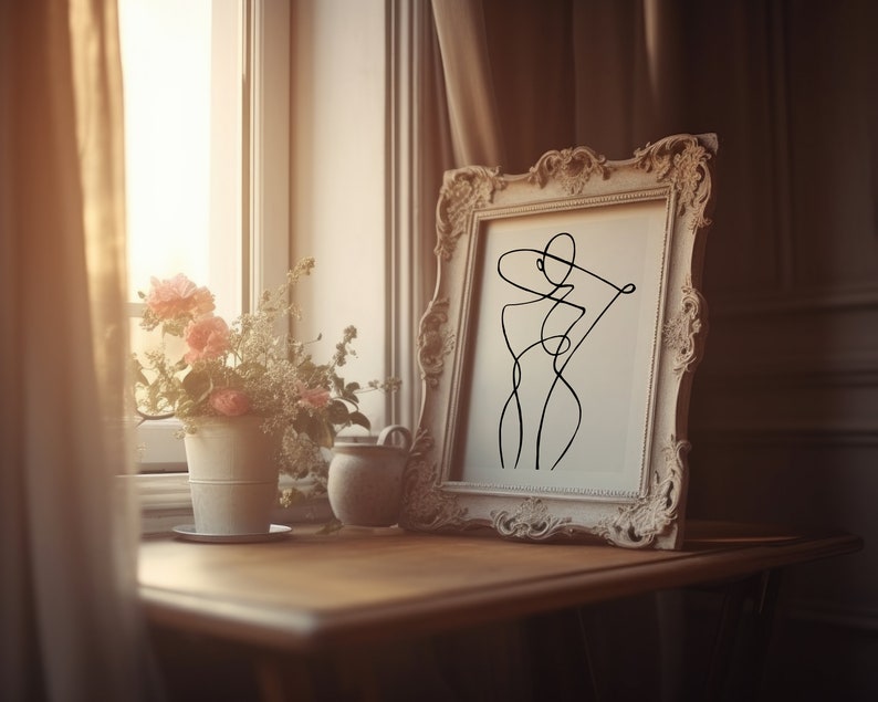 K&ouml;nnte beinhalten: Ein wei&szlig; gerahmtes Bild mit einer schwarzen Linienzeichnung einer Person mit Hut. Das Bild steht auf einem Holztisch mit einer Vase mit rosa Blumen und einem kleinen Keramikgef&auml;&szlig;.