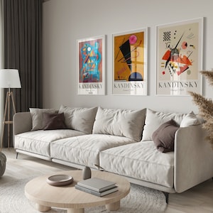 May include: Three framed abstract art prints by Wassily Kandinsky, featuring geometric shapes and vibrant colors, hanging on a white wall above a white sofa with brown throw pillows.