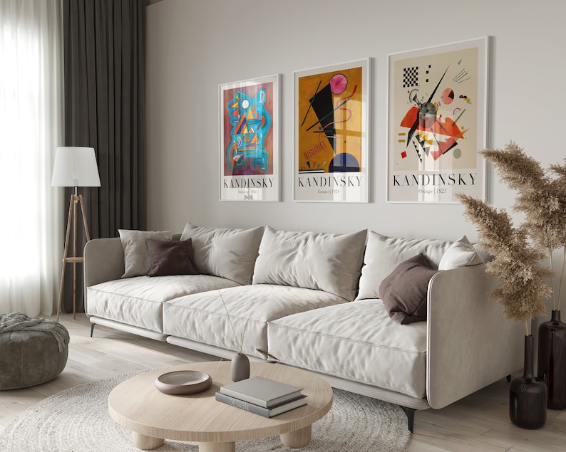 May include: Three framed abstract art prints by Wassily Kandinsky, featuring geometric shapes and vibrant colors, hanging on a white wall above a white sofa with brown throw pillows.