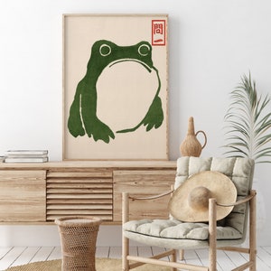May include: A framed art print featuring a green frog illustration on a beige background. The frog has a large open mouth and simple lines. A red Japanese character is in the upper right corner. The artwork is displayed in a light wood frame.