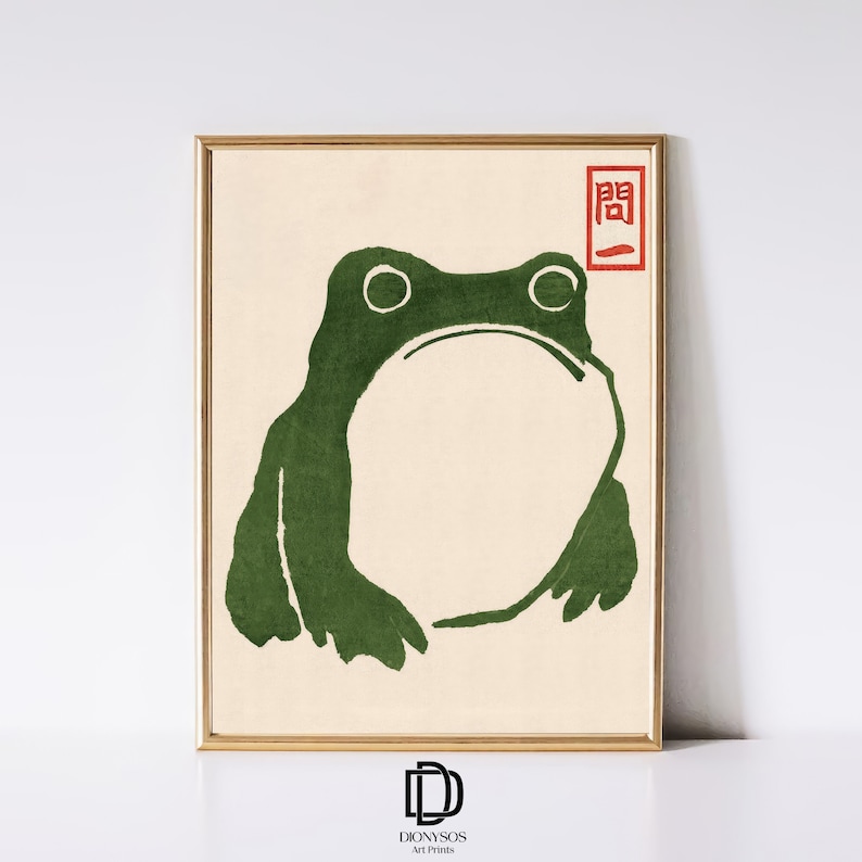 May include: A framed art print featuring a green frog illustration in a minimalist style. The frog has a large, open mouth and is set against a cream-colored background. A red Japanese character is in the upper right corner. The frame is gold.