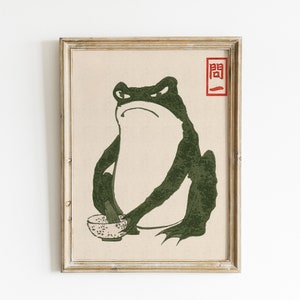 May include: A framed print of a green frog holding a bowl with a black and white pattern. The frog has a serious expression and is sitting on a beige background. The print has a red stamp with the Japanese character "関" in the top right corner.