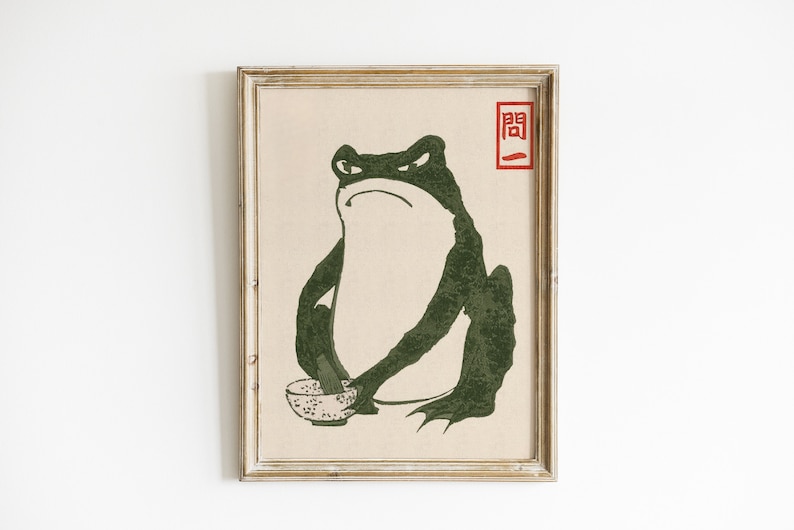 May include: A framed print of a green frog holding a bowl with a black and white pattern. The frog has a serious expression and is sitting on a beige background. The print has a red stamp with the Japanese character "関" in the top right corner.
