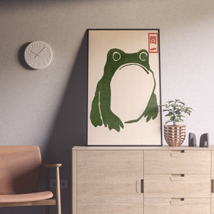 May include: A framed art print featuring a green frog illustration with Japanese text in the upper right corner. The artwork is displayed on a light-colored cabinet, with a small plant and decorative items. A round clock hangs on the wall.