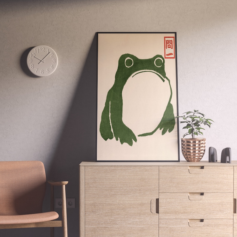 May include: A framed art print featuring a green frog illustration with Japanese text in the upper right corner. The artwork is displayed on a light-colored cabinet, with a small plant and decorative items. A round clock hangs on the wall.