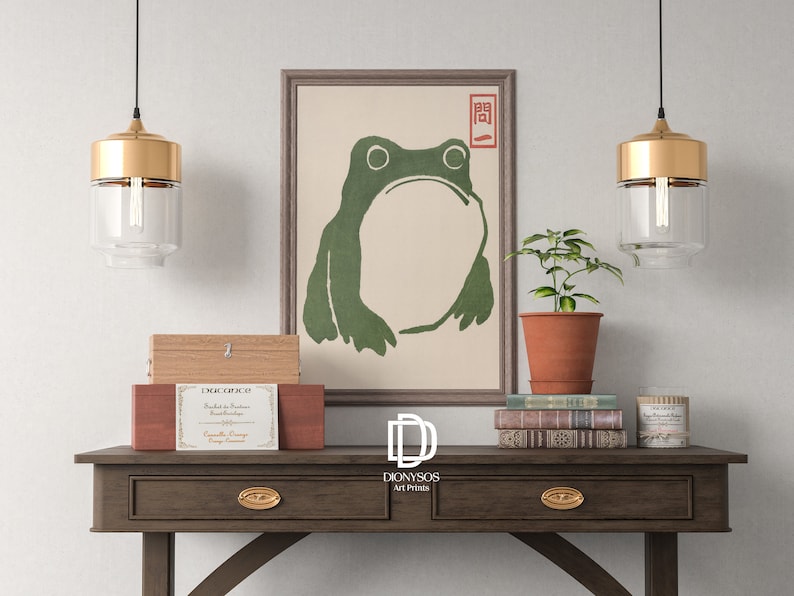 May include: A framed print of a green frog with a minimalist design hangs above a wooden console table. The table holds decorative boxes, books, and a potted plant. Two pendant lights with gold accents hang above the table.