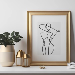 K&ouml;nnte beinhalten: Ein goldfarbener gerahmter Druck mit einer schwarzen Linienzeichnung einer stilisierten menschlichen Figur. Der Druck befindet sich auf einem wei&szlig;en Regal mit einer goldenen Vase und zwei goldenen Kerzenhaltern.