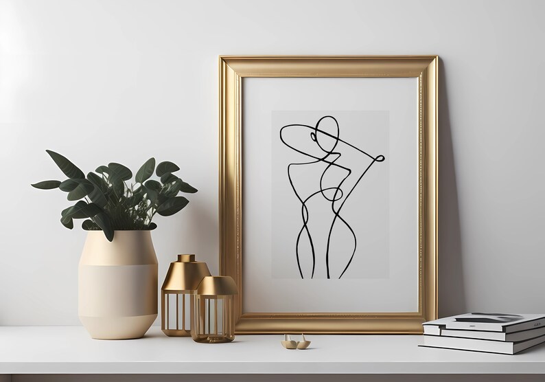 K&ouml;nnte beinhalten: Ein goldfarbener gerahmter Druck mit einer schwarzen Linienzeichnung einer stilisierten menschlichen Figur. Der Druck befindet sich auf einem wei&szlig;en Regal mit einer goldenen Vase und zwei goldenen Kerzenhaltern.