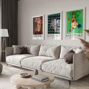 May include: Three framed art prints featuring abstract geometric designs by Wassily Kandinsky. The prints are in white frames and feature the artist's name and the title and date of each work. The prints are hanging on a white wall above a white sofa.