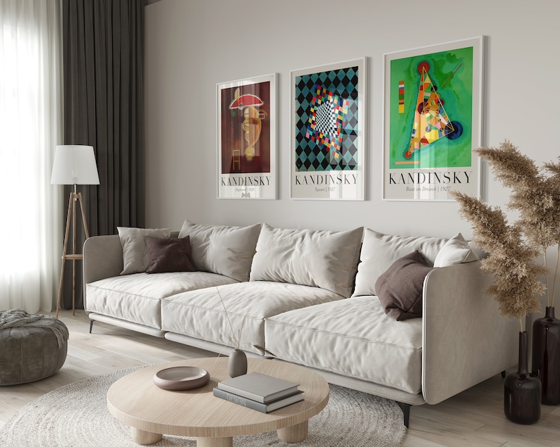 May include: Three framed art prints featuring abstract geometric designs by Wassily Kandinsky. The prints are in white frames and feature the artist's name and the title and date of each work. The prints are hanging on a white wall above a white sofa.