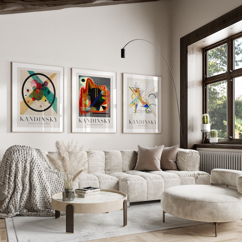 May include: Three framed abstract art prints by Wassily Kandinsky, featuring geometric shapes and vibrant colors, hanging on a white wall above a light beige sofa with a throw blanket and pillows.