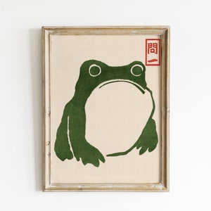 May include: A framed print featuring a stylized green frog illustration. The frog has large eyes and a wide mouth, set against a beige background. A red Japanese character is in the upper right corner.