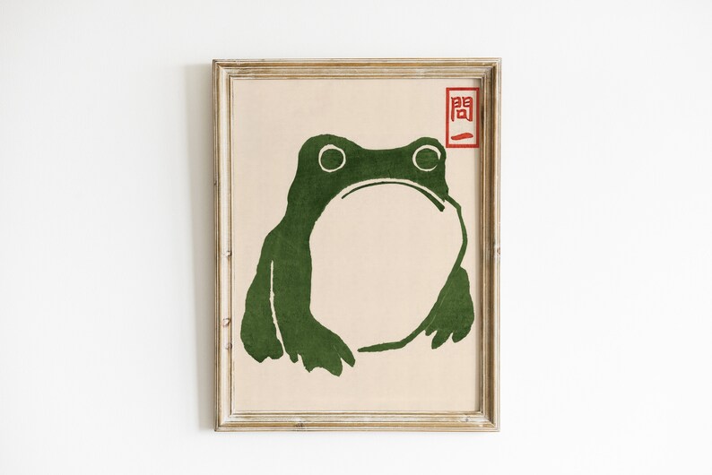May include: A framed print featuring a stylized green frog illustration. The frog has large eyes and a wide mouth, set against a beige background. A red Japanese character is in the upper right corner.
