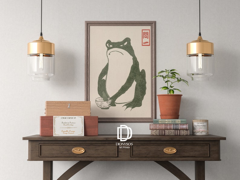 May include: A framed print of a green frog with a bowl in its hands. The print is on a wooden table with a wooden box, a potted plant, and three books. The print has the text "関" in red.