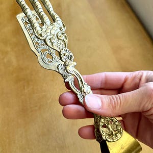 May include: A brass serving tong featuring a three-pronged claw design at the top, with intricate embossed scrollwork and a bird motif on the handle. The metal has a muted gold tone with a textured finish, and the handle ends in a smooth loop.