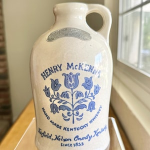 May include: A white ceramic jug with a blue floral design and the text "Henry McKenna Hand Made Kentucky Whiskey Fairfield, Nelson County Kentucky Since 1853".