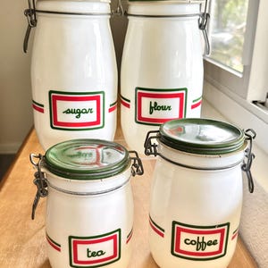 May include: A set of four white glass storage canisters with green metal lids and wire bail closures. Each canister features a rectangular label with red and green borders, printed with the words sugar, flour, tea, and coffee in cursive script.