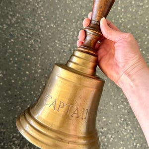 May include: A vintage brass bell with a wooden handle. The bell has the word "CAPTAIN" engraved on its side. The handle is a dark brown colour, and the bell itself has a golden hue. The bell is held in a hand.