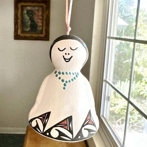 Puede incluir: Un adorno de muñeca de cerámica blanca con cabeza negra y cara sonriente. La muñeca tiene un collar turquesa y un dobladillo estampado con diseños negros, rojos y blancos. Cuelga de una cinta rosa.