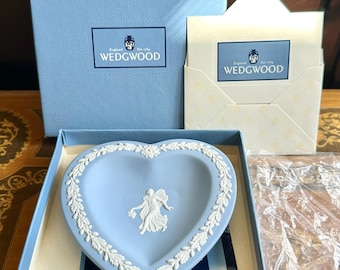 May include: A light blue heart-shaped Wedgwood jasperware dish featuring a white relief of a figure and a floral border. The dish is presented in a light blue box with a matching envelope, both displaying the Wedgwood logo and text.