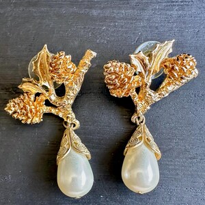 May include: A pair of gold-toned earrings featuring a branch design with pinecones and pear-shaped faux pearls. The earrings have a post back and are set against a dark background.