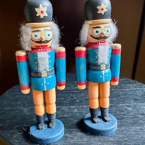 May include: Two wooden nutcracker figurines standing on circular blue bases. Each figure wears a black hat with a gold star, a blue jacket with red accents, and tan trousers with black boots. They feature white hair, painted faces, and mustaches.